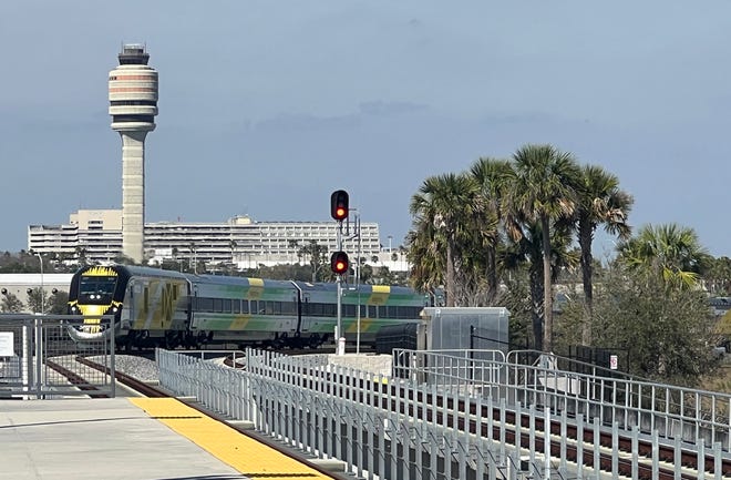 A Brightline train rolls at Orlando International Airport with the air traffic control tower in the background.