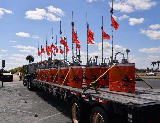 A truckload of mystery aquatic items are sitting on a tractor trailer bed in the parking lot of Patrick Space Force Base. The bright orange objects are numbered, have antenna and each one has two boat propellers protruding from them.